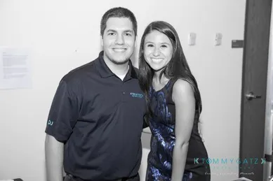 A man and woman pose together, smiling. The man wears a black polo shirt and the woman wears a patterned dress.