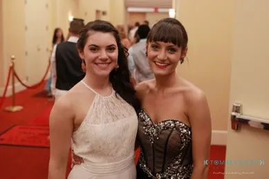 Two women in formal dresses smile for a photo on a red carpet with people in the background.