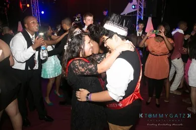 Two teenagers wearing crowns dance closely at a party, surrounded by other people in formal attire.