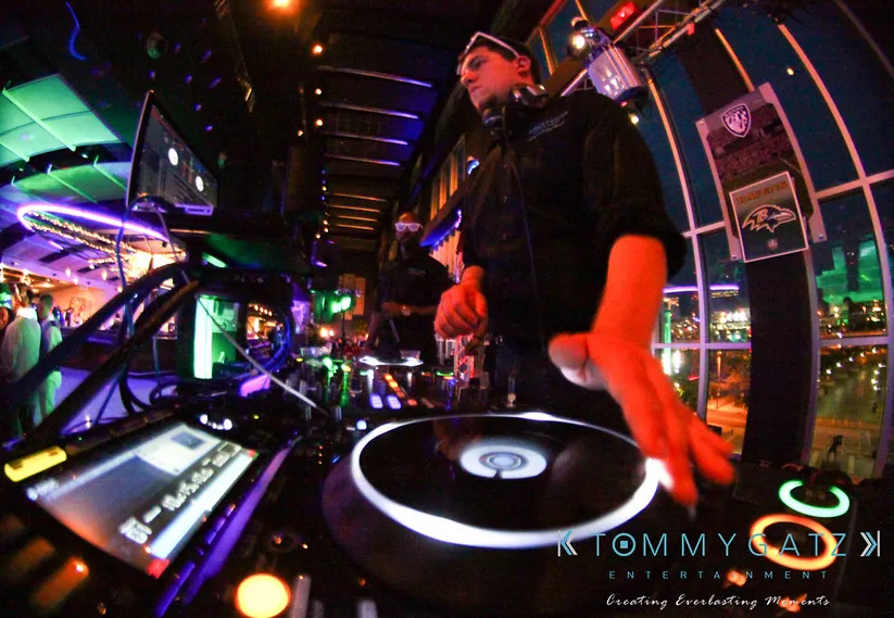 A DJ wearing a headset plays music at a nightclub, surrounded by colorful lights and sound equipment.