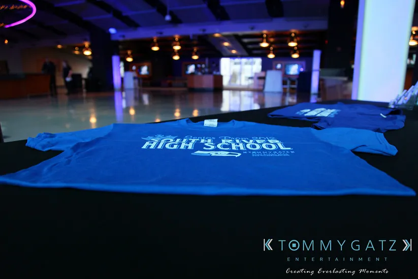 A blue t-shirt featuring the text "Dulaney High School" is displayed on a table in a dimly lit room. The logo "Tommy Gatz Entertainment" is visible in the corner.