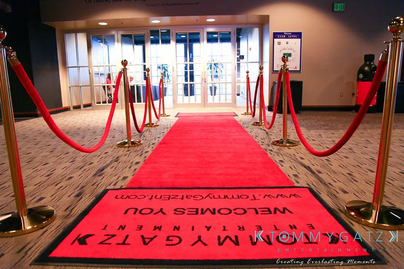 A red carpet with stanchions leads to glass doors. A welcome mat with "Tommy Gatz Entertainment" is on the floor. The background has a poster and a trash can.