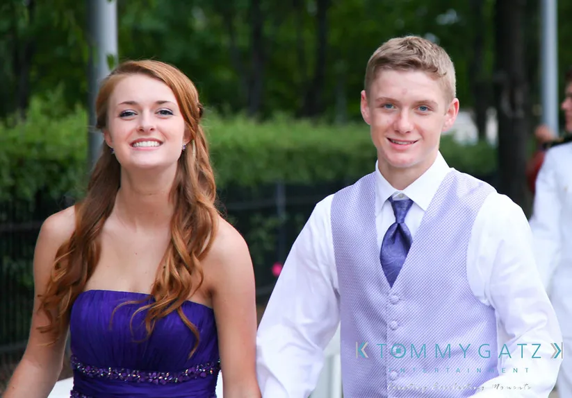 A young woman in a purple dress and a young man in a white shirt and purple vest pose outdoors.