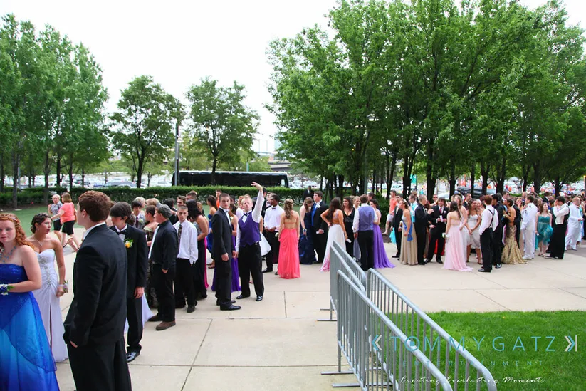 A large group of people in formal attire is gathered outdoors, forming a line near metal barriers. Trees are visible in the background.