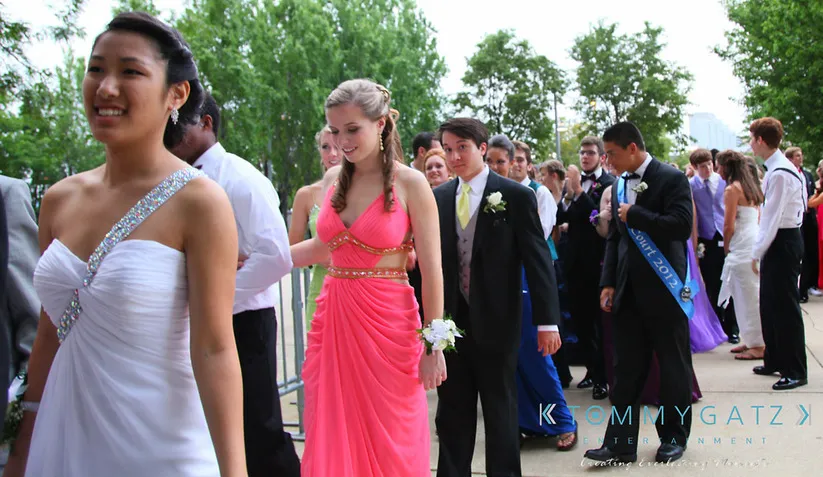 A group of young adults in formal attire walk in a line, possibly at a prom event. Students appear happy and are dressed in colorful gowns and suits. Trees and a building are visible in the background.