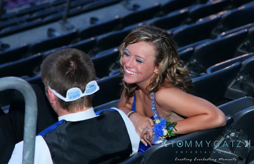 A young woman in a blue dress smiles while talking to a man in formal attire with a cap. They are seated in a stadium.