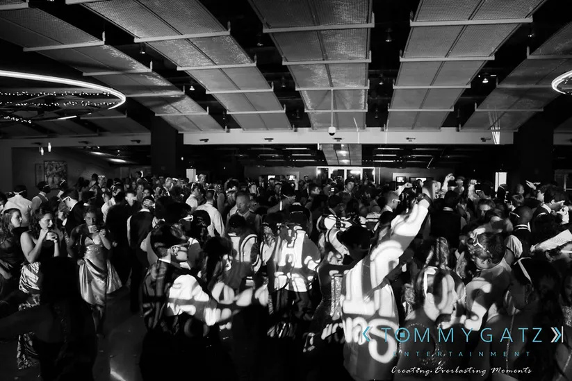 A large crowd dances in a dimly lit venue with Tommy Gatz Entertainment signage projected on the floor.