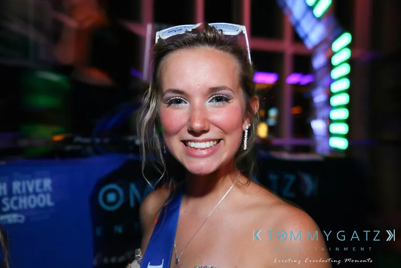 A person smiling at an event, wearing a sash and sunglasses on their head, with colorful lights in the background.
