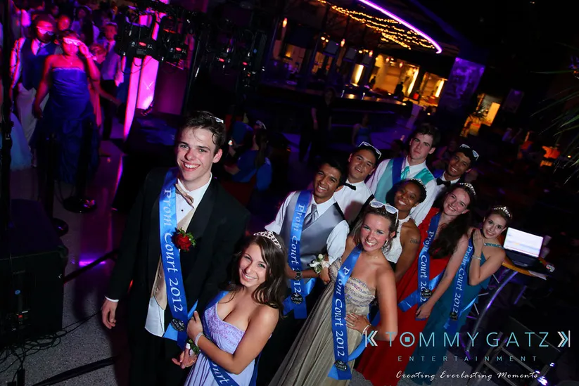 A group of smiling people dressed in formal attire, wearing sashes labeled "Prom Court 2012," pose in a decorated venue with colorful lighting.