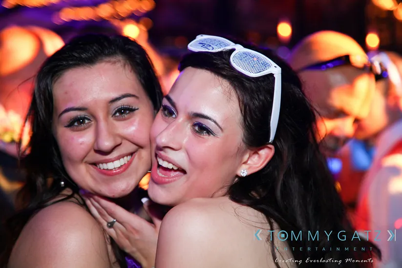Two people smile at a lively party. One wears white sunglasses on their head. Bright, colorful lights create a festive atmosphere.
