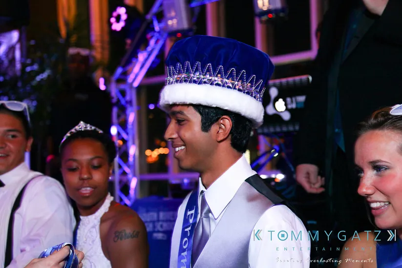 A young man wearing a royal blue crown and sash stands at a prom event, smiling. Other attendees are visible in the background.