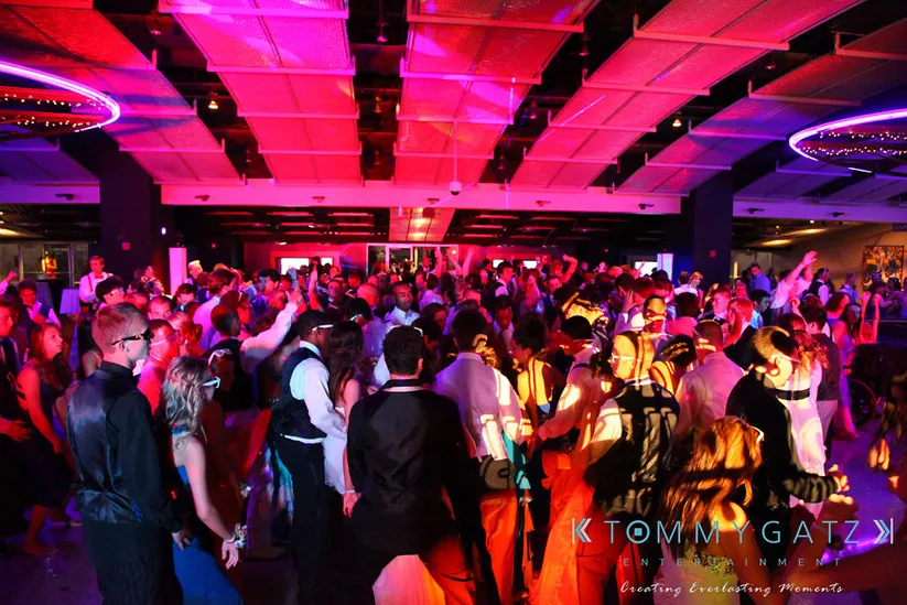 A large crowd of people dancing in a dimly lit, colorful event space with vibrant overhead lighting.