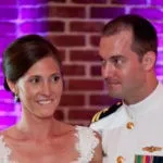 A couple stands together in front of a brick wall, with the man in a military uniform and the woman in a white dress.