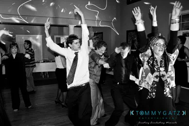 A group of people dancing energetically in a room with a wooden floor, party decorations, and a buffet table in the background.