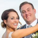 A bride and groom smile at the camera, with the bride wearing a white dress and the groom in a suit and tie.