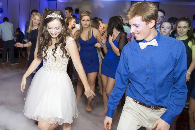 A young couple dances together in a room filled with people, surrounded by fog. The woman wears a white dress, and the man wears a blue shirt with a bow tie.