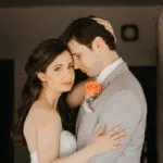 A couple embraces closely, with the man in a gray suit and the woman in a white dress. An orange boutonniere is on the man's lapel.