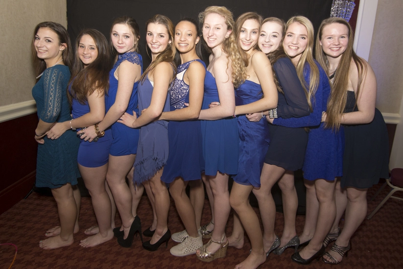 A group of ten people wearing blue dresses stand closely together in a line, smiling at the camera.