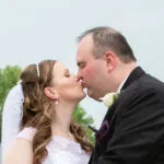 A bride and groom kiss outdoors, with trees in the background.