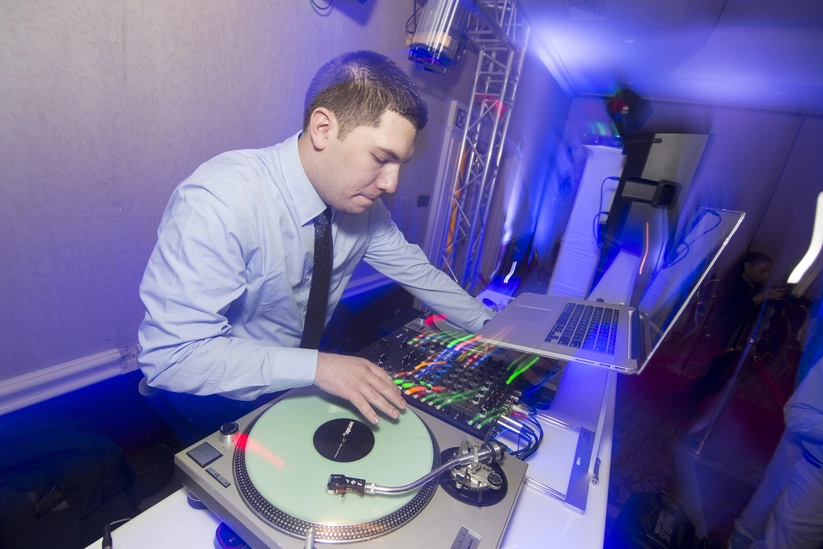 A DJ in a blue shirt is mixing tracks on a turntable and mixer setup, surrounded by vibrant blue lights at an indoor event.