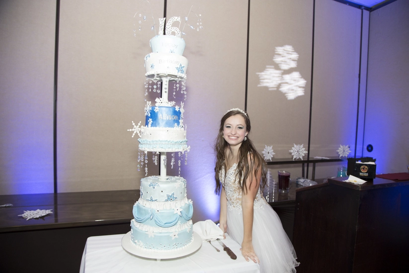 A young woman in a white dress stands beside a tall blue and white tiered cake decorated with snowflakes and the number 16 on top.