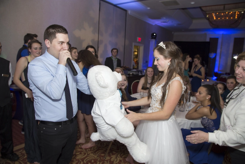 A man in a blue shirt holds a microphone while a woman in a white dress holds a large white teddy bear at a social event with several people in the background.