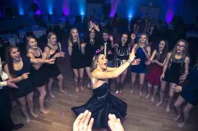 A group of young women in dresses are gathered in a circle on a dance floor, clapping and cheering. The room is dimly lit with blue and purple lights.