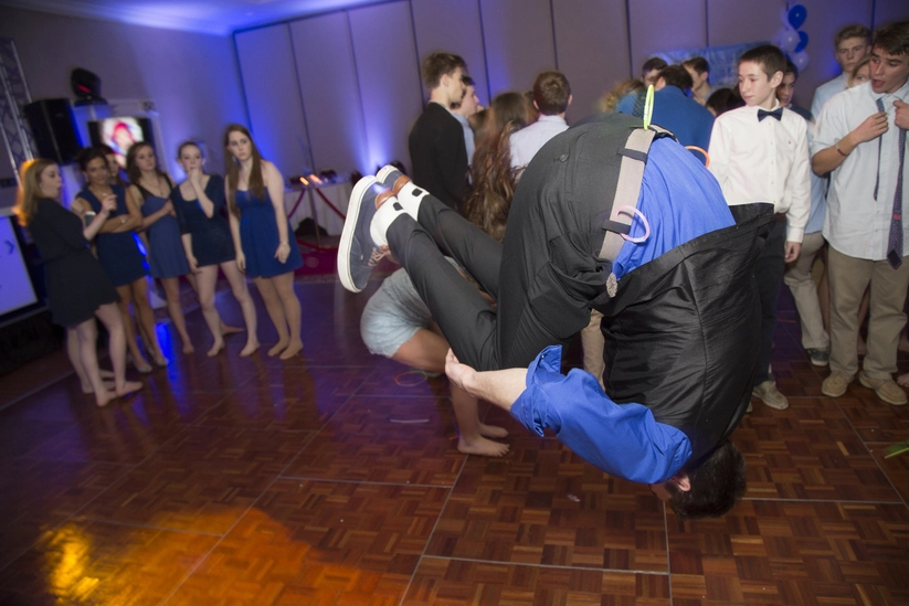 A person wearing a blue shirt performs a backflip on a dance floor, surrounded by onlookers in formal attire at a party.