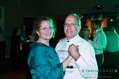 A couple is dancing together at an indoor event. They are smiling and surrounded by other people on the dimly lit dance floor.