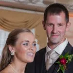 A couple in formal attire poses indoors; the woman wears earrings and a necklace, while the man has a boutonniere on his suit.