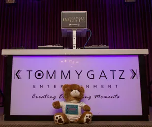 DJ booth with Tommy Gatz Entertainment logo, a laptop on top, and a stuffed lion sitting in front.