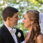 A bride and groom smile at each other outdoors, surrounded by greenery.