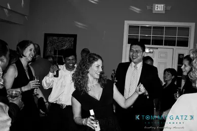 A group of people in formal attire are smiling and dancing at a party. Some hold drinks. The setting appears to be indoors with a closed door and EXIT sign in the background.