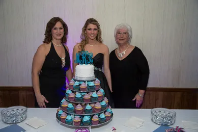 Three women stand behind a table with a cupcake tower and a small cake on top, decorated with a blue topper.