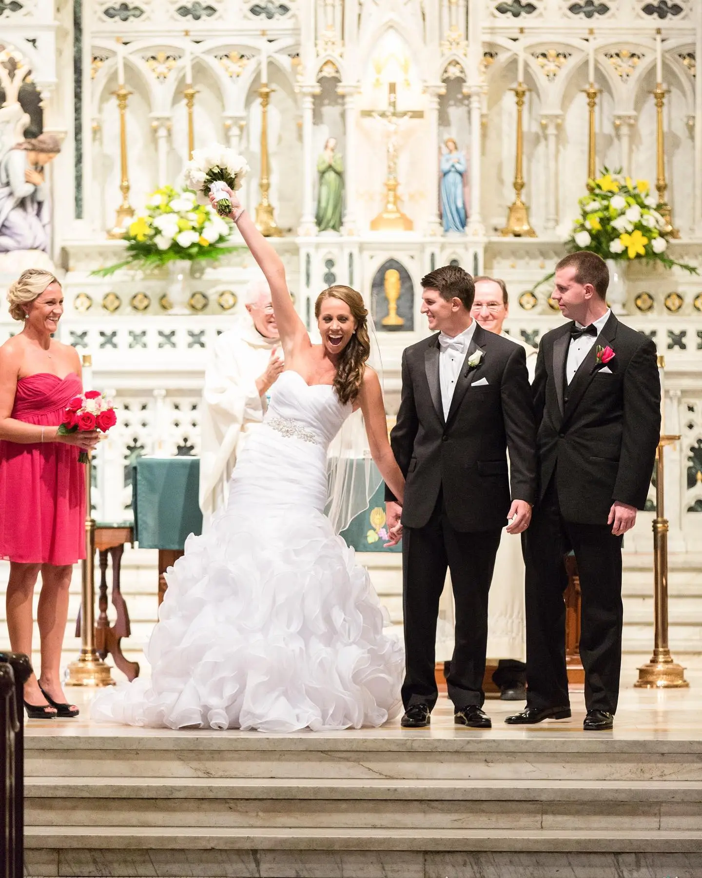 A bride in a white dress raises her bouquet while standing with the groom and two attendants in a church setting.