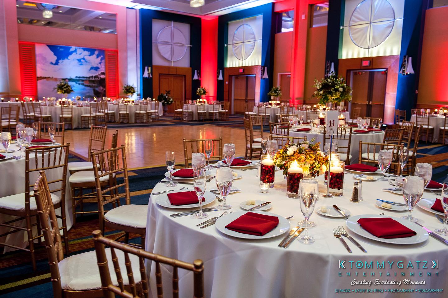 A decorated banquet hall with round tables set for an event. Tables have red napkins, candles, and floral centerpieces. Elegant lighting and large wall art enhance the room's ambiance.