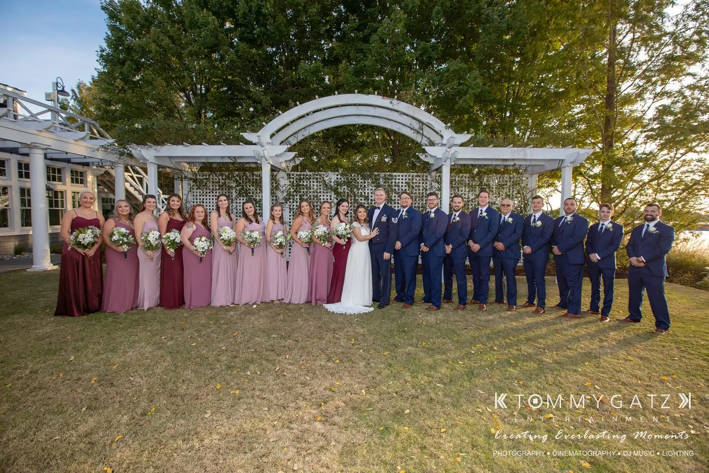 A large wedding party stands in a garden setting, featuring bridesmaids in pink dresses and groomsmen in navy suits, with greenery and a wooden archway in the background.