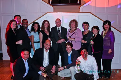 A group of people posing together indoors, dressed in formal and semi-formal attire, some holding drinks. They stand and sit in front of a staircase.