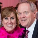 A smiling man and woman are posing together at an event. The woman wears a pink feather boa, and the man is in a suit and tie.