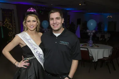 A woman wearing a sash and tiara stands next to a man in a black shirt. They are in a decorated room with tables and balloons.