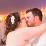 A couple kissing at a wedding, with the woman wearing a white dress and hair accessory. The background shows soft lighting.