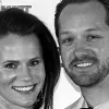 A grayscale photo of a smiling woman and man standing side by side against a white background.