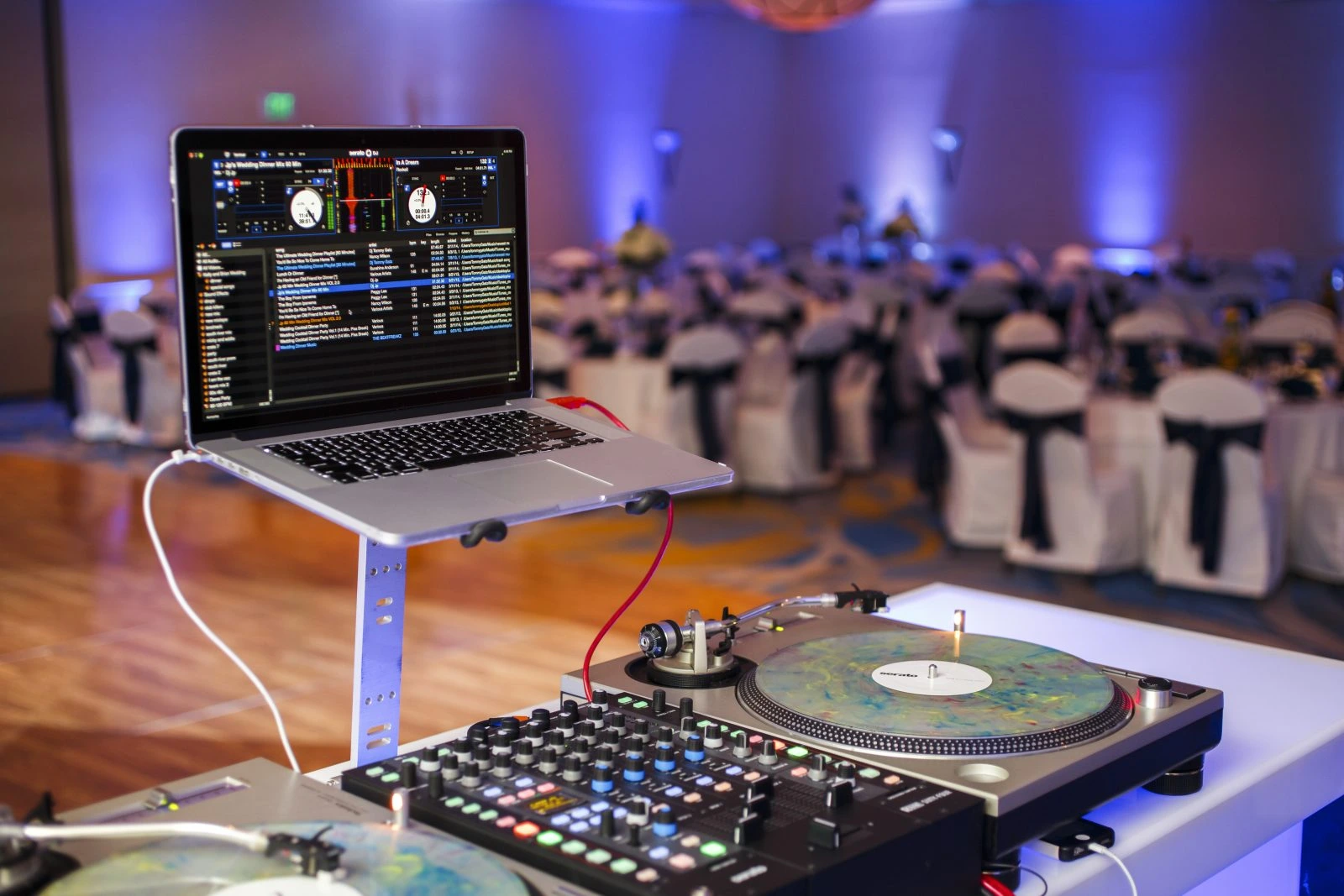 DJ setup with a laptop, turntables, and mixer in the foreground. Background shows a decorated banquet hall with tables and chairs.