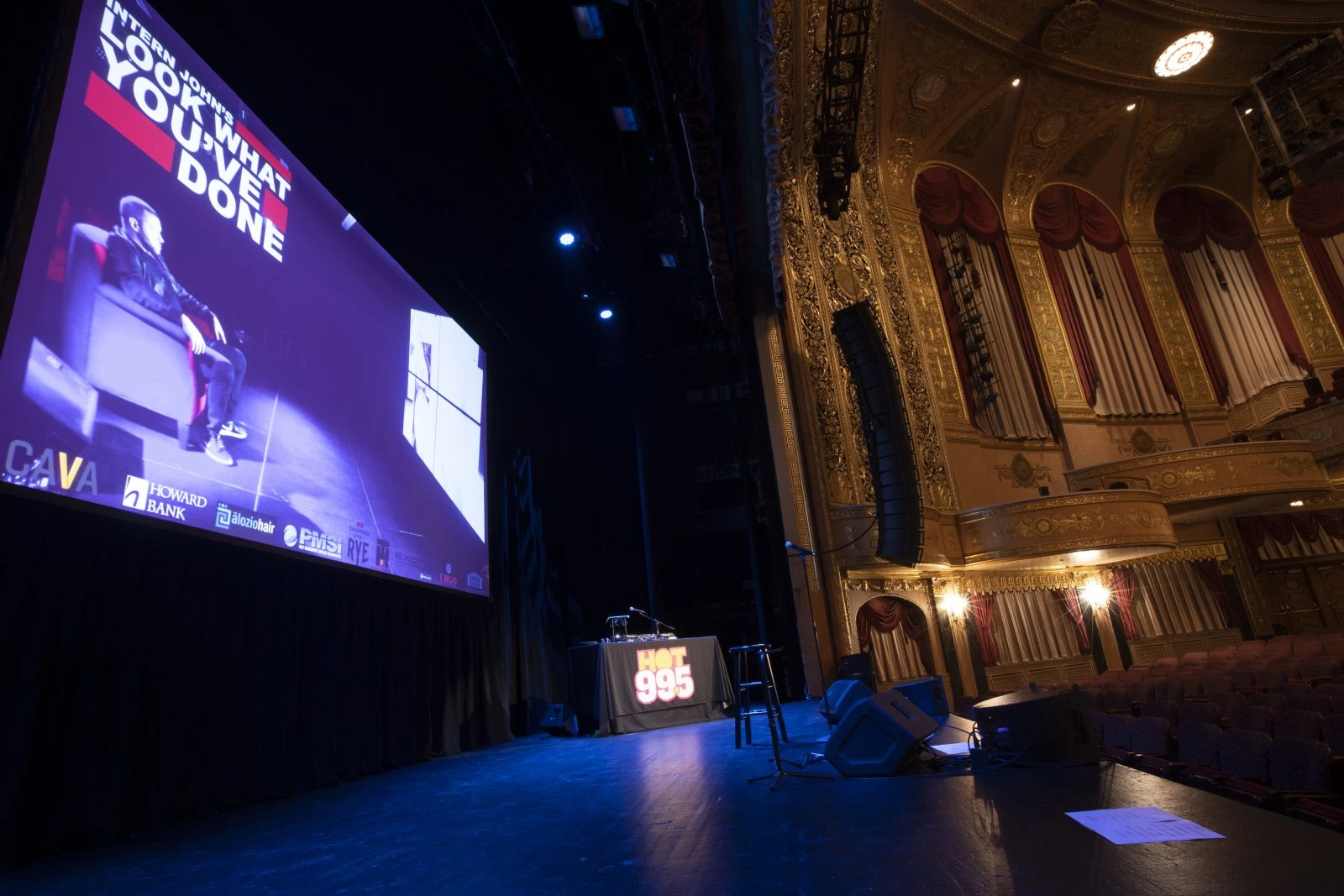 An empty stage with a screen showing a promotional image next to a table and microphone, in a large ornate theater.