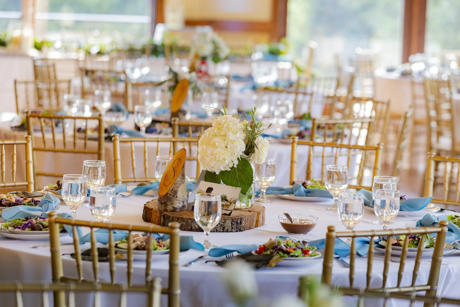 A wedding reception setup with round tables covered in white tablecloths, gold chairs, floral centerpieces, place settings, and glasses of water arranged neatly.