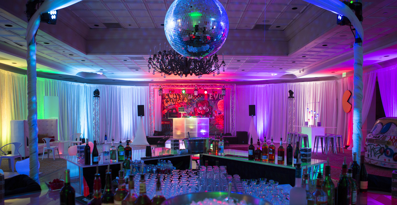 A vibrant nightclub interior with a disco ball ceiling, colorful lights, a bar with bottles and glasses, and lounge seating on the sides.
