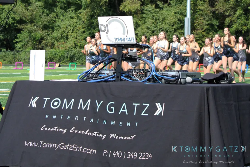 DJ setup on a table with "Tommy Gatz Entertainment" branding. Cheerleaders performing in the background on a sports field.