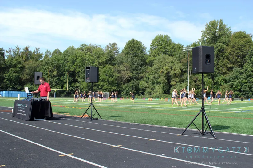 DJ playing music on a sports field with dancers practicing in the background. Speakers are set up on either side of the DJ table.