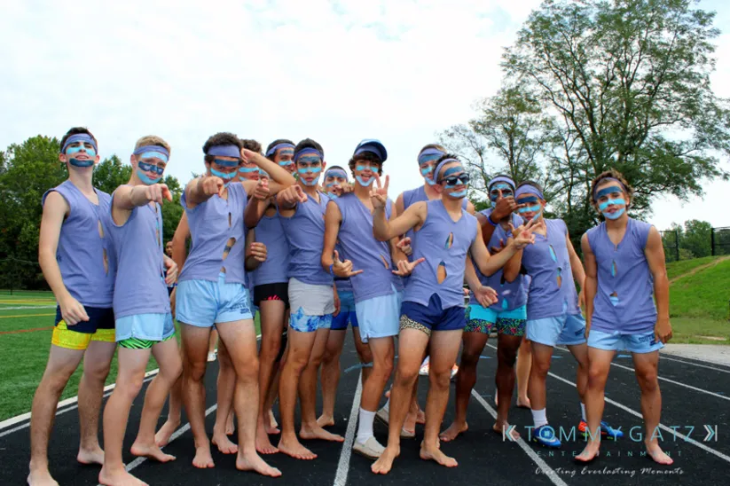 A group of people wearing matching purple tank tops and face paint pose on a running track. Trees are visible in the background.