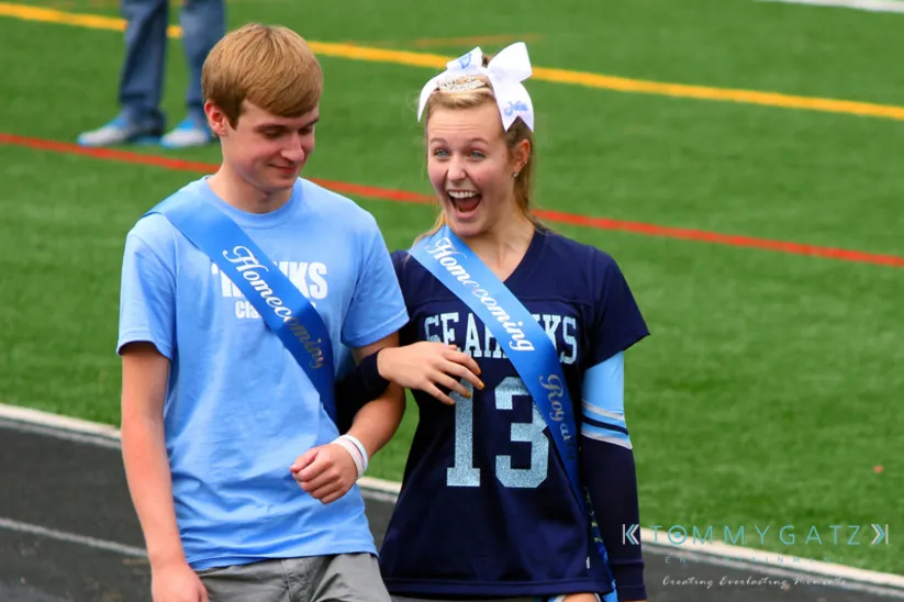 Two students wearing homecoming sashes walk on a sports field. The person on the right looks amused, wearing a bow and jersey.